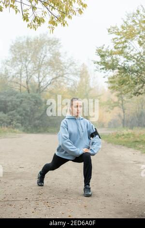 Ragazza attiva in auricolari, felpa con cappuccio blu, leggini neri e sneakers che fanno esercizio per allungare le gambe sul percorso forestale contro gli alberi al mattino Foto Stock