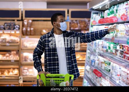 Giovane uomo nero in maschera protettiva che acquista prodotti caseari a. supermercato durante la quarantena del coronavirus Foto Stock