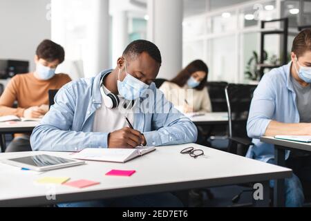 Studente di sesso maschile nero in maschera seduto alla scrivania e di scrittura Foto Stock