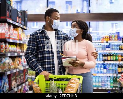 Millennial coppia nera in maschere protettive che controllano la loro lista settimanale di acquisto del cibo, che comprano i prodotti al centro commerciale Foto Stock