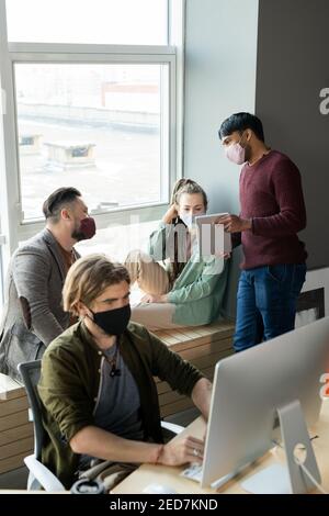 Tre giovani designer o manager in casualwear e maschere protettive discussione delle informazioni online sul display del tablet tenuto da uno di loro Foto Stock