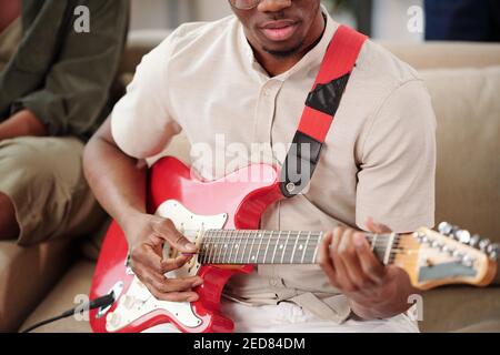 Giovane africano in casualwear che suona la chitarra elettrica mentre si siede sul divano contro uno dei suoi amici durante la riunione a. a casa per la festa Foto Stock