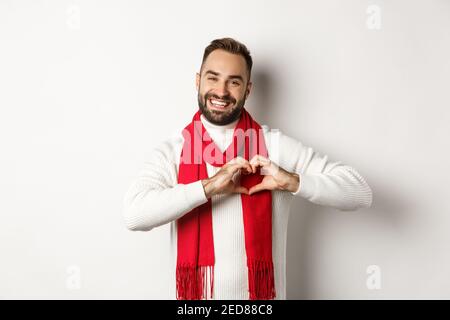 Vacanze di Natale e concetto di Capodanno. Felice padre che mostra il segno del cuore e sorridente, ti amo gesto, indossando maglione invernale e sciarpa, bianco Foto Stock