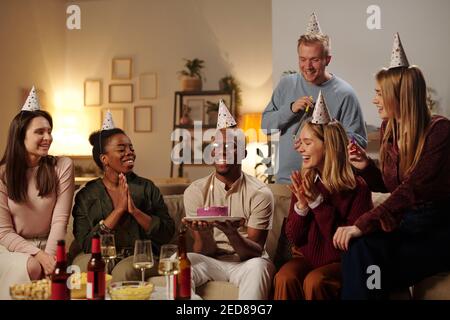Felici giovani amici interculturali congratulandosi con un ragazzo africano sorridente per il suo compleanno e aggrappando le mani mentre si guarda una torta deliziosa Foto Stock