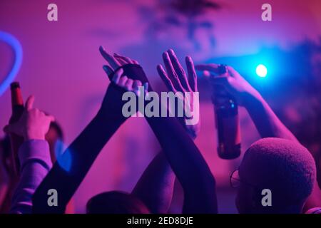 Alzò le braccia di giovani amici interculturali che ballano a casa festeggiamenti e bevande contro le luci rosa durante il fine settimana Foto Stock