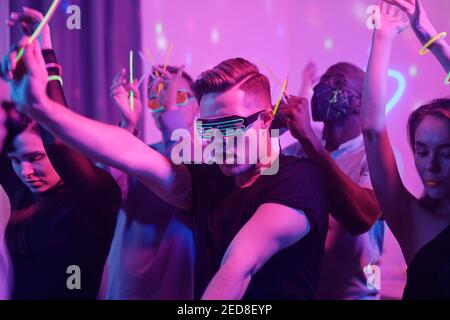 Giovane eccitato uomo in casualwear e discoteca occhiali e suo amici interculturali con bastoni incandescenti che ballano insieme mentre si godono a casa festa Foto Stock
