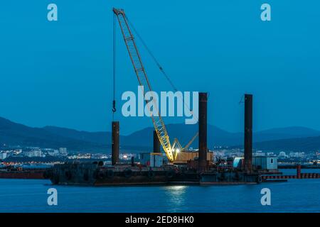 Gru cargo che lavora in piccoli porti, costruzione di porti industriali o banchine in serata, trasporto di merci marine e concetto di sviluppo logistico. Foto Stock