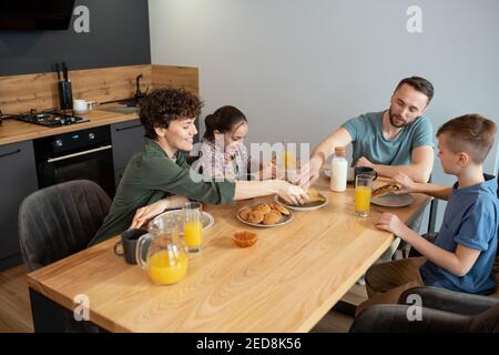 Felice famiglia di quattro seduti al tavolo servito nel cucina e gustando la colazione mentre padre e madre prendono due ultimi panini fatti in casa Foto Stock