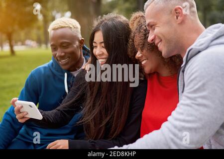 Multi etnia amici esterni guardare lo schermo dello smartphone. Gruppo diverso gente afro americano asiatico caucasico passare il tempo insieme multirazziale maschio femmina studente seduto panca parco all'aperto Foto Stock