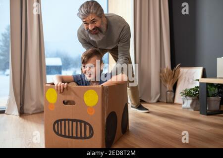 Felice uomo maturo bearded in casualwear spingendo scatola di cartone con suo nipote mentre entrambi si divertono durante il gioco in salotto Foto Stock