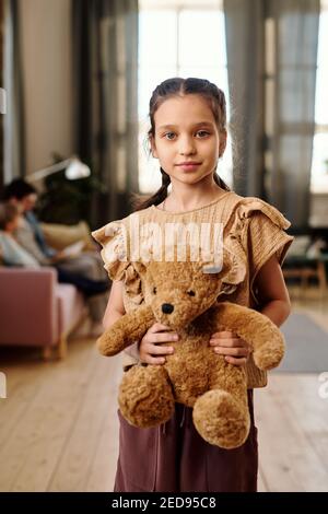 Bambina graziosa con il teddybear marrone morbido che li guarda mentre si sta davanti alla fotocamera contro la sua famiglia seduto sul divano in soggiorno Foto Stock