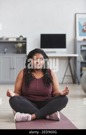 Ritratto verticale di una donna afro-americana curvy meditating a casa mentre si siede sul tappetino e si sta godendo di yoga a casa pratica Foto Stock