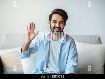 Saluto. Amico uomo orientale che si fa ciao mentre si siede sul divano a casa Foto Stock