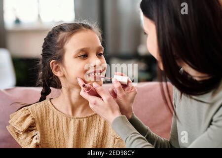 Carina bambina in abito seduta sul divano di fronte di sua madre che aiuta la figlia con lucentezza del labbro mentre preparazione per l'evento di celebrazione Foto Stock