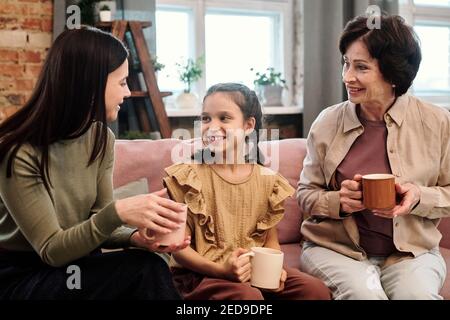 Due donne felici contemporanee in casualwear e adorabile bambina con bevande calde seduti sul divano in soggiorno e avere discussione Foto Stock