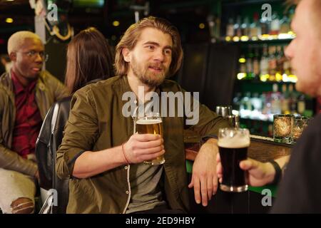 Giovane uomo bearded in casualwear che ha birra dal banco del bar mentre si guarda il suo amico di fronte alla fotocamera e. parlandogli Foto Stock