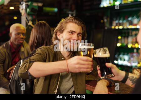 Felice giovane uomo sopportato in casualwear guardando il suo amico mentre si è aggraffati con bicchieri di birra davanti alla fotocamera e parlare Foto Stock