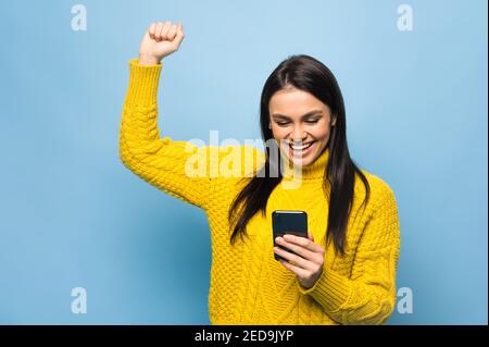 Ritratto di una giovane donna caucasica felice, tiene smartphone a portata di mano, gioisce di una buona notizia o ha ricevuto un messaggio inaspettato, si trova su sfondo blu isolato Foto Stock