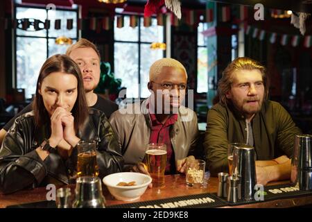 Gruppo di giovani amici interculturali in casualwear in piedi presso il bar bancone con bevande e spuntini mentre si guarda la trasmissione di calcio in pub Foto Stock