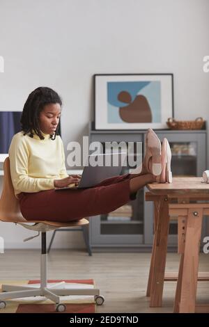 Verticale ritratto a tutta lunghezza di giovane donna afroamericana usando il laptop a casa con i piedi sul tavolo, spazio di copia Foto Stock