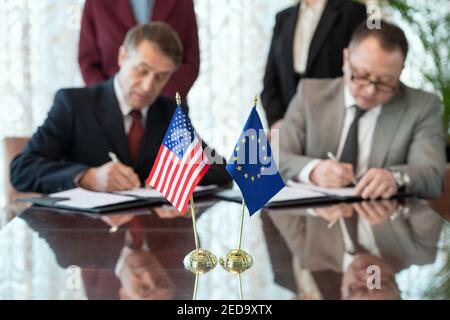 Bandiere degli Stati Uniti e dell'Unione europea sul tavolo contro due partecipanti firmano il contratto dopo la negoziazione e la conclusione dell'accordo Foto Stock
