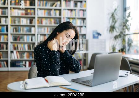 Una donna sesaurita manager, freelance o studentessa in eyeglase siedono al tavolo, utilizzando il laptop, sviluppare un progetto. Stanco caucasico lavoro eccessivo donna d'affari, avendo un teso e mal di schiena, hanno bisogno di un riposo Foto Stock