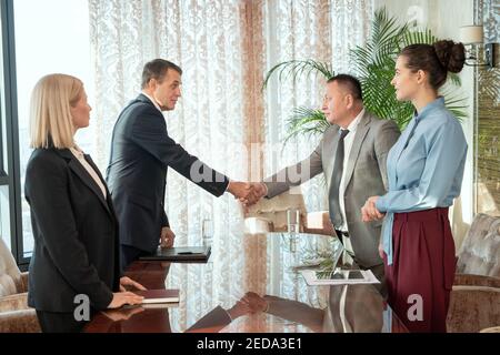 Uomini d'affari maturi di successo o delegati in formalwear salutarsi l'un l'altro con handshake sopra il tavolo dopo la negoziazione e la conclusione di un accordo Foto Stock