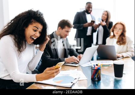 Gioiosa dipendente seduto alla scrivania, guarda il telefono e si rallegra della fortuna. Donna d'affari afroamericana che legge le buone notizie, felice circa il progetto riuscito o l'avvio, lavoro ben fatto Foto Stock