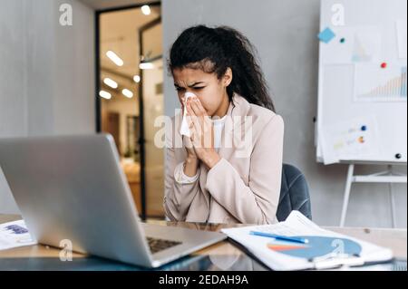 Donna d'affari siede alla scrivania, usando carta tissue, starnutendo. Bella donna dipendente sensazione di malessere, malata con naso che corre. Donna afro-americana che lavora o studia a distanza, mentre la malattia Foto Stock