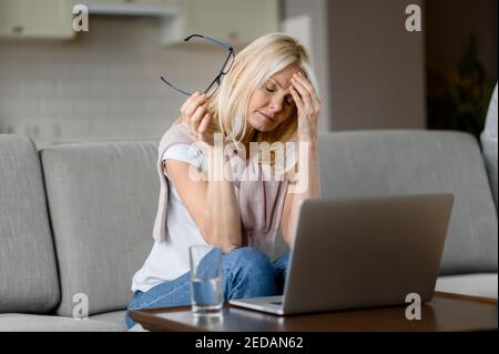 Triste caucasica metà età freelance femminile, stanco di lavorare straordinario online, seduto a casa in soggiorno, tolto i suoi occhiali, chiuso gli occhi, ha bisogno di un riposo Foto Stock