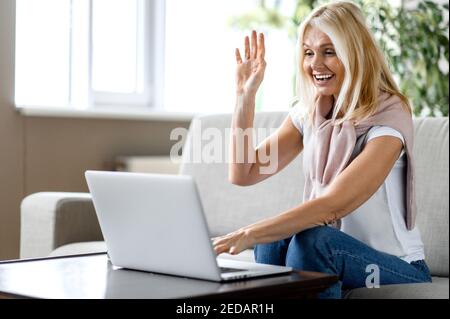 Sorridente anziana caucasica con occhiali seduti sul divano in soggiorno, utilizzando il computer portatile, mentre lavora a distanza, comunica con colleghi di lavoro o amici, onde mano, saluti Foto Stock