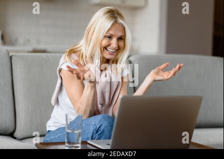 Felice sorridente donna caucasica anziana con occhiali seduti sul divano in soggiorno, utilizzando il computer portatile, mentre lavora a distanza, comunica con i colleghi di lavoro Foto Stock