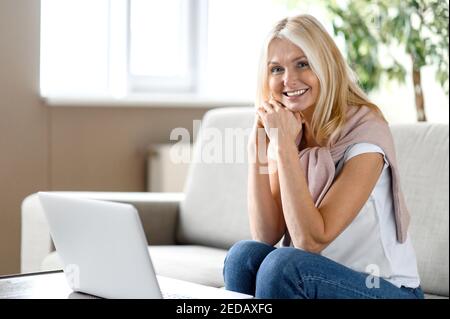 Rilassata bionda caucasica di mezza età si siede a casa in soggiorno, utilizzando il computer portatile, guarda la fotocamera e sorridendo felice, lavoro o studio a distanza Foto Stock