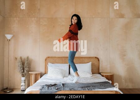 Allegra e rilassante donna etnica asiatica in maglione sorridente e saltellante a letto mentre ci si diverte a casa Foto Stock