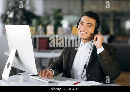 Giovane adulto maschio sta consultando i clienti, lavoro nel servizio di supporto. Felice uomo ispanico seduto alla scrivania, indossando le cuffie, funziona come operatore di call center, comunicando con i clienti Foto Stock