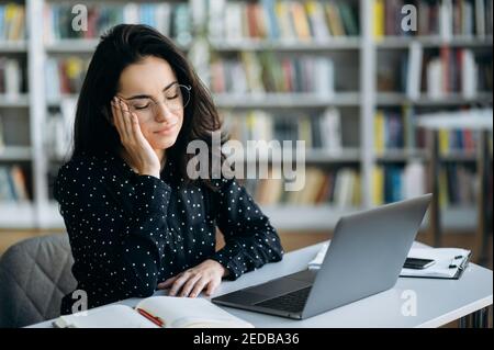 Una giovane donna adulta si siede al banco di lavoro in ufficio, avendo un terribile mal di testa. Donna di affari esausta in occhiali sul lavoro teso, sentendosi stanco, overwork, bisogno di una rottura Foto Stock