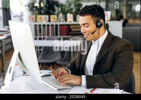 Soddisfatto uomo d'affari ispanico, manager o tutor seduto al banco di lavoro indossando le cuffie chattando online con colleghi o studenti e sorridendo Foto Stock