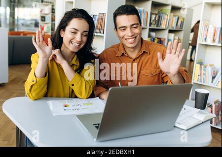 Colleghi o studenti multirazziali soddisfatti, seduti sul posto di lavoro, comunicano tramite videochiamata con colleghi o amici sorridenti e guarda la fotocamera del notebook e le mani sventolanti Foto Stock