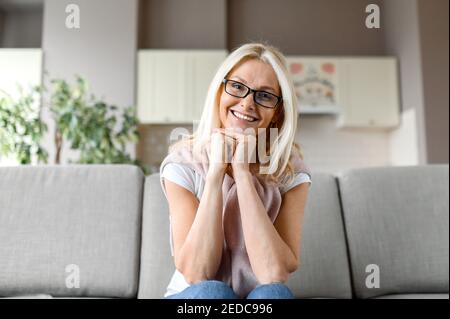 Ritratto di affascinante donna caucasica matura indossando occhiali, seduto sul divano in soggiorno, guardando direttamente la macchina fotografica con un sorriso amichevole Foto Stock