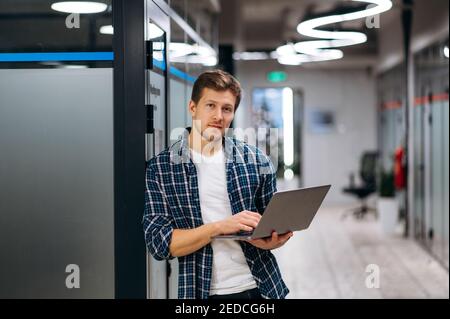 Stand freelance di successo in un ufficio moderno, utilizzando un computer portatile. Un uomo d'affari sicuro e attraente che lavora sul proprio progetto o startup. Giovane adulto maschio dipendente guardare direttamente la fotocamera e sorridere Foto Stock