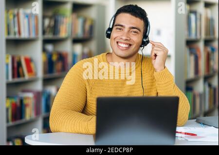 Un consulente di sesso maschile sorridente seduto alla scrivania, indossando le cuffie. Il tipo ispanico lavora come operatore del call center. Il giovane adulto sta parlando con i clienti, concetto di servizio di supporto Foto Stock