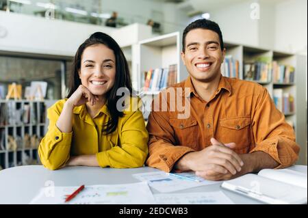 Colleghi o studenti multirazziali soddisfatti, seduti in interni, parlano in videochiamata con colleghi o amici sorridenti e guarda la fotocamera Foto Stock