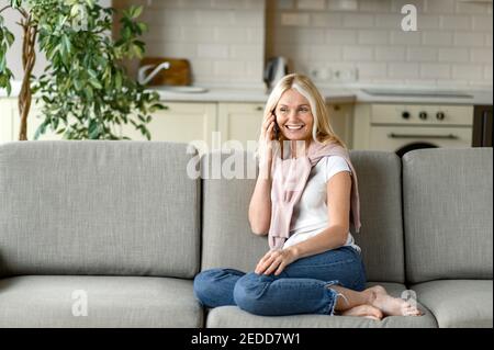 Una donna sorridente di mezza età usa lo smartphone, parla con amici o familiari. Senior donna bionda in abiti casual si siede sul divano con telefono cellulare, parla e sorridente, concetto di comunicazione Foto Stock