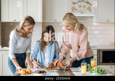 Tre generazioni di donne trascorrono il tempo insieme in cucina, insegnate alla bambina a tagliare l'insalata, sorridendo e felici di stare insieme Foto Stock