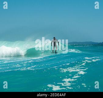 Un uomo surfs su un aliscafo surf sulle onde turchesi a Mangawhai Heads, Nuova Zelanda. Cielo blu chiaro. Foto Stock