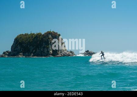 Un uomo surfs su un aliscafo surf sulle onde turchesi a Mangawhai Heads, Nuova Zelanda. Cielo blu chiaro. Foto Stock