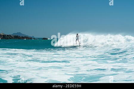 Un uomo surfs su un aliscafo surf sulle onde turchesi a Mangawhai Heads, Nuova Zelanda. Cielo blu chiaro. Foto Stock