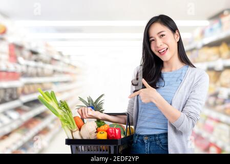 Bella donna asiatica tenendo carrello di shopping pieno di verdure e. generi alimentari nel supermercato Foto Stock