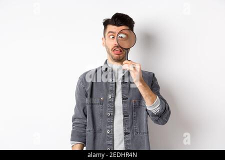 Divertente giovane uomo che squinting, mostrando volti con lente d'ingrandimento, in piedi su sfondo bianco Foto Stock