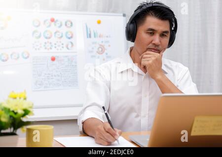 Uomo d'affari asiatico che parla con il team tramite videoconferenza scrivere nota su carta. Uomini d'affari che usano il laptop e le cuffie per le riunioni online. Uomo che usa Foto Stock